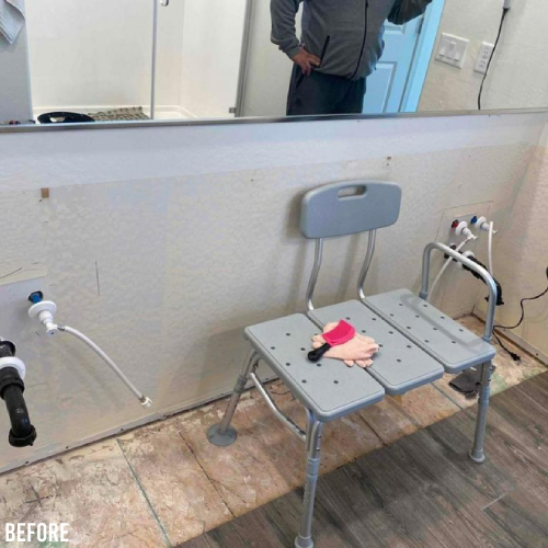 A bathroom under renovation shows exposed pipes and an unfinished floor. A gray shower chair with a backrest is in front of the sink area. A mirror is above the sink space, reflecting a person.