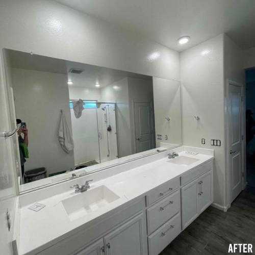 Renovated bathroom featuring a large mirror, double sinks with white cabinetry, bright lighting, and a visible shower area in the background.