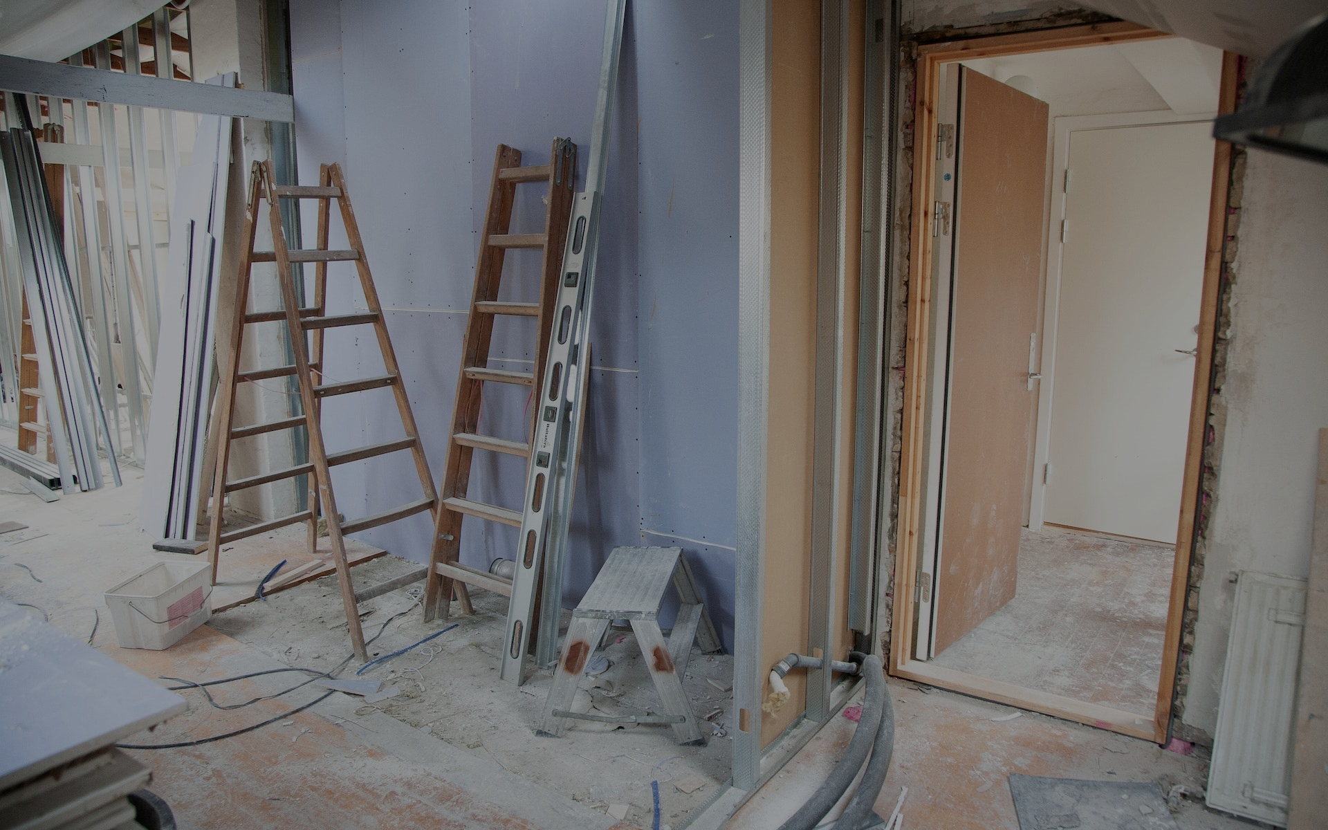Interior under renovation with ladders, drywall, and construction materials scattered. Walls partially constructed, wires and tools visible, open door leads to another room.